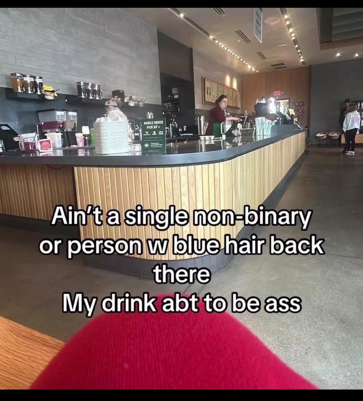 The meme is a photo of a coffee shop, but with a humorous and satirical twist. The text overlayed on the image reads: "Ain't a single non-binary or person with blue hair back there / My drink is abt to be ass". The image is likely meant to be humorous and ironic, poking fun at the idea that someone is being judged or looked down upon for their appearance or identity.